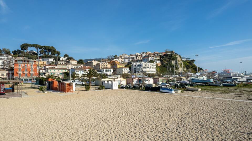 A coastal town with buildings on a hillside, a sandy beach, and boats docked along the shore.