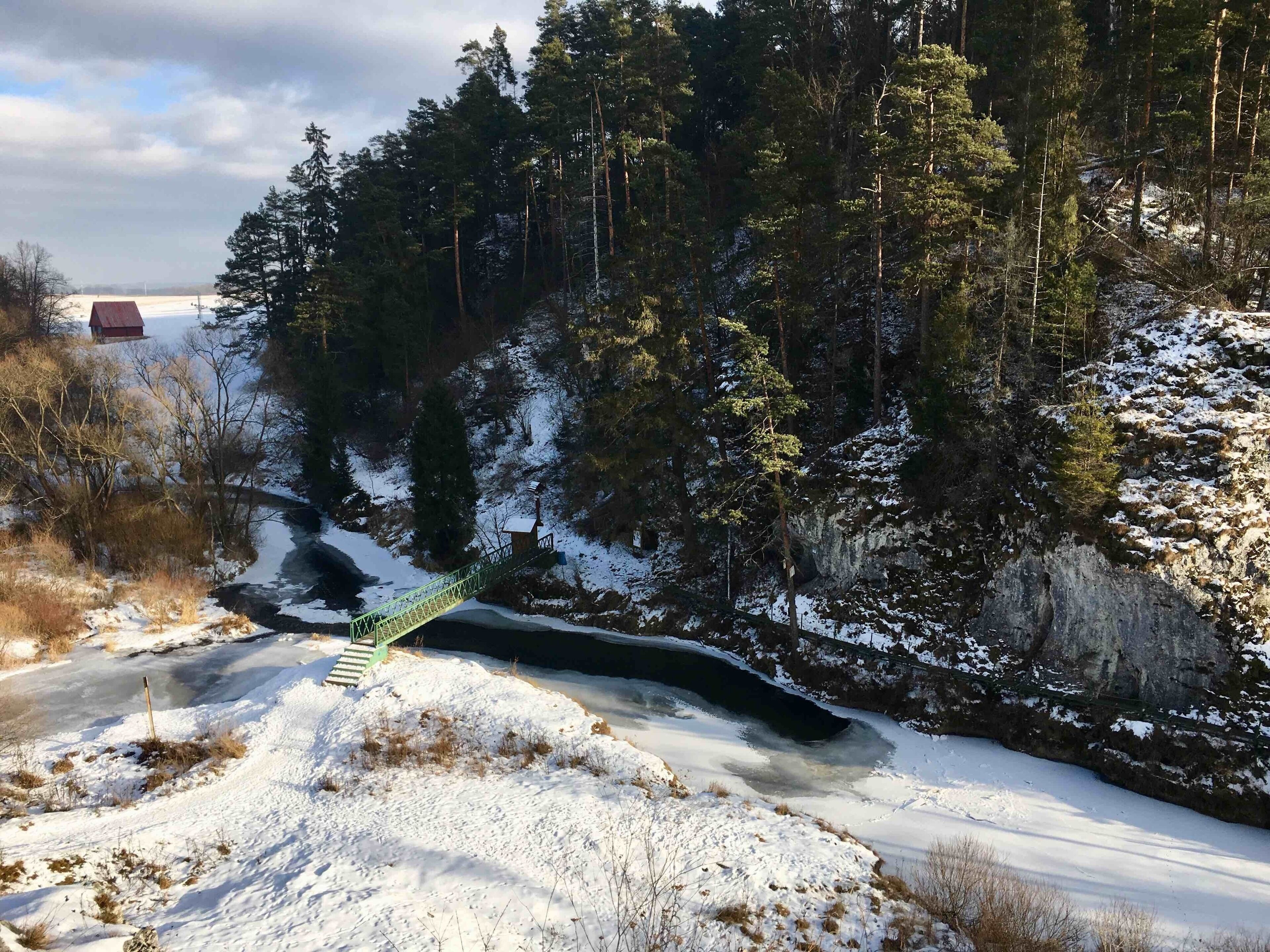 A snow-covered landscape with a wooden bridge over a frozen river, surrounded by dense forest and a distant red building.