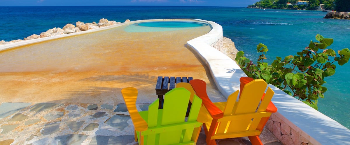 A pool with a curved edge, surrounded by a stone walkway. There are two colorful Adirondack chairs near the pool.