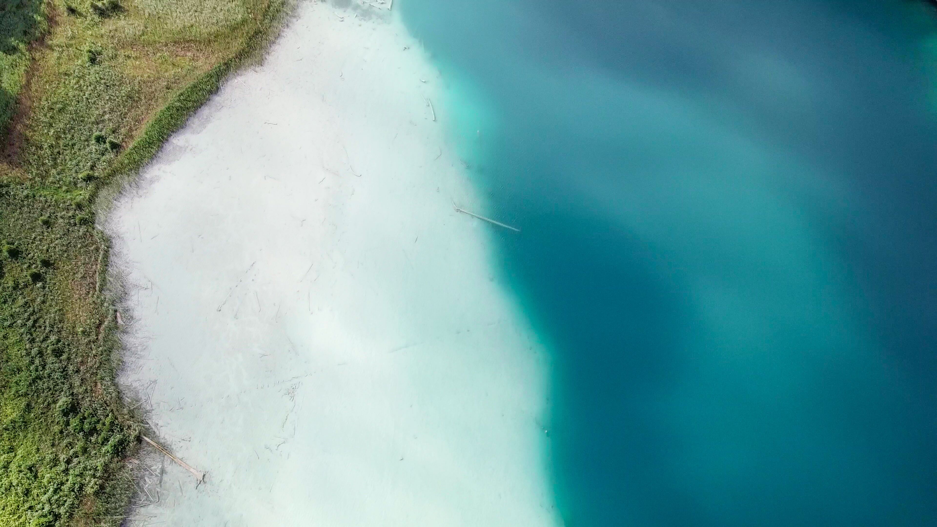 A top down, drone capture of Weissensee lake in Austrian Alps. The water changes color from white, turquoise to navy blue. Green shore of the lake. A bit of overcast. Serenity and peacefulness