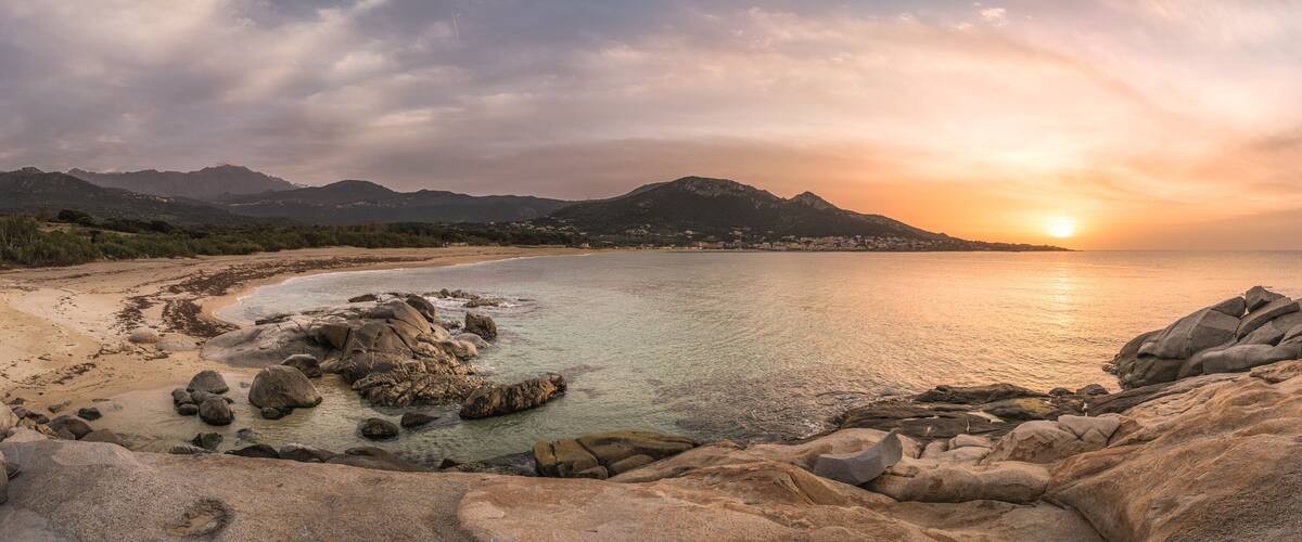 A coastal landscape with a sunset, rocky shoreline, and distant mountains.