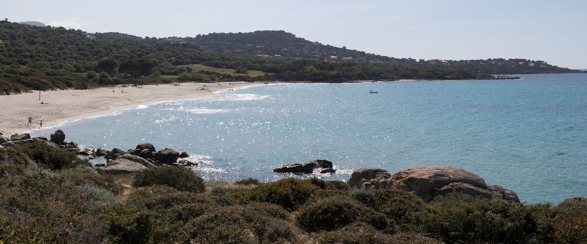 A coastal landscape with a sandy beach, rocky outcrops, and a hilly backdrop.