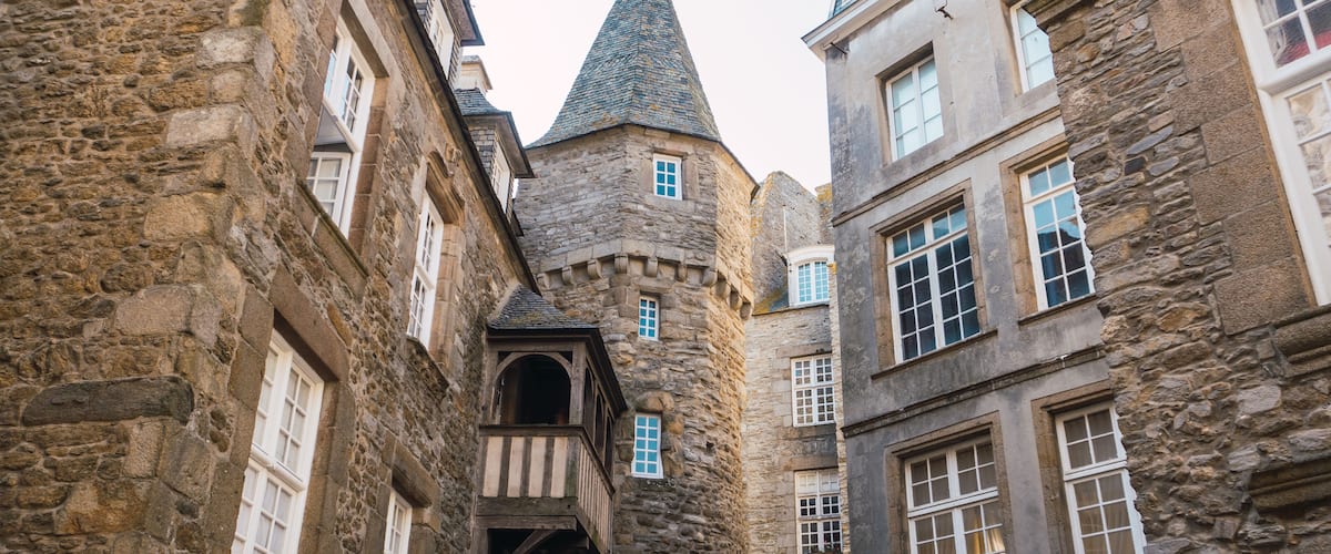 A narrow medieval street with stone buildings, a tower, and a wooden balcony.
