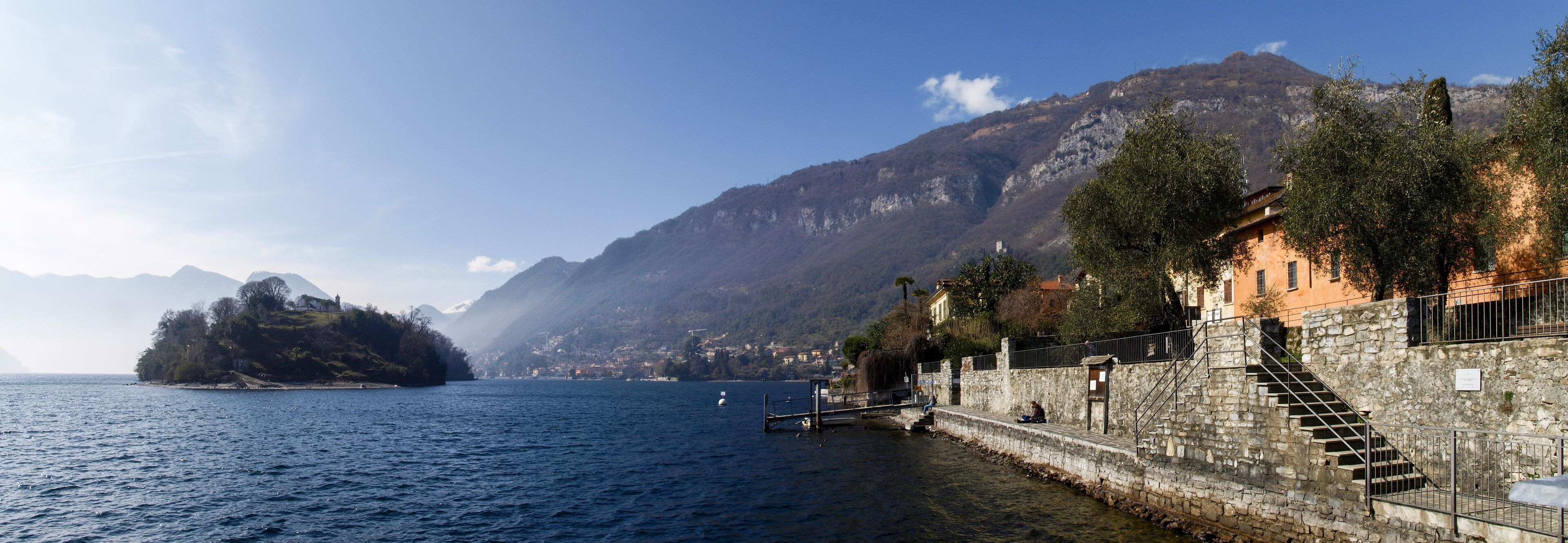 A coastal town with a stone staircase leading to a waterfront, a small island in the background, and mountains.