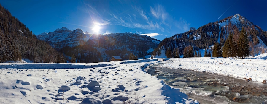 A snowy mountain landscape with a clear sky, a frozen river, and a wooden cabin.