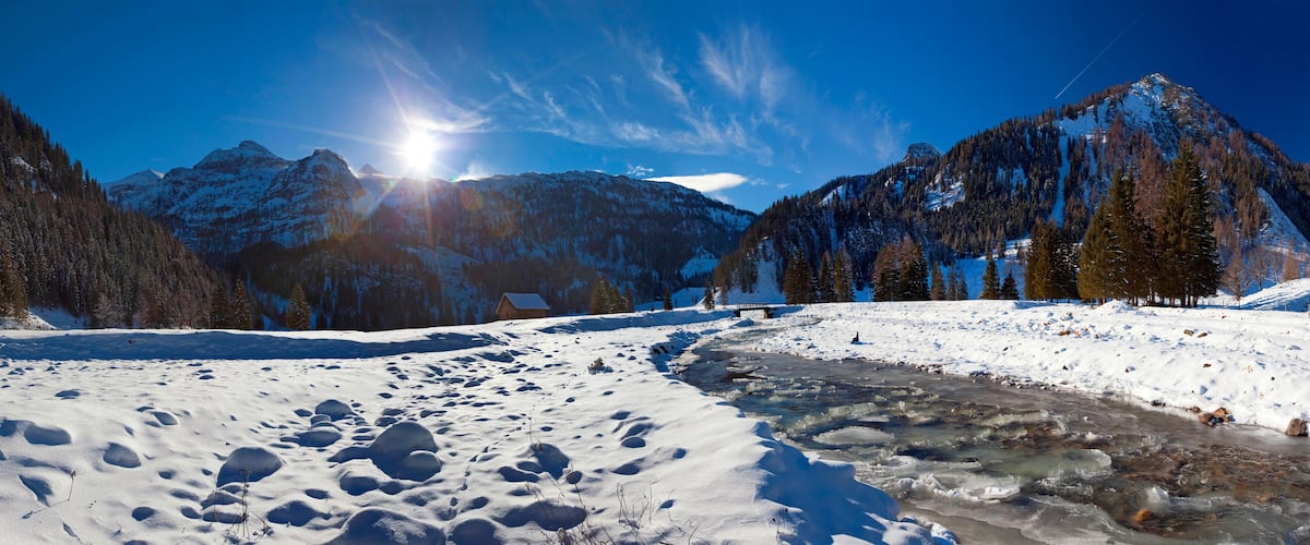 A snowy mountain landscape with a clear sky, a frozen river, and a wooden cabin.
