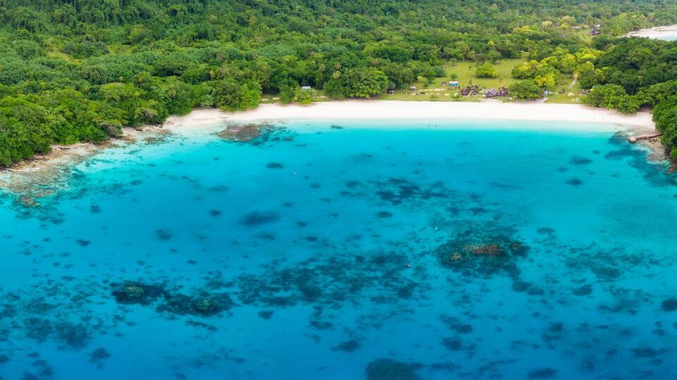 An aerial view of a tropical island with a white sandy beach, turquoise waters, and dense green forest.