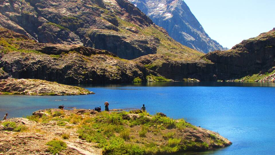 A mountainous landscape with a clear blue lake, rocky terrain, and a few people near the water's edge.
