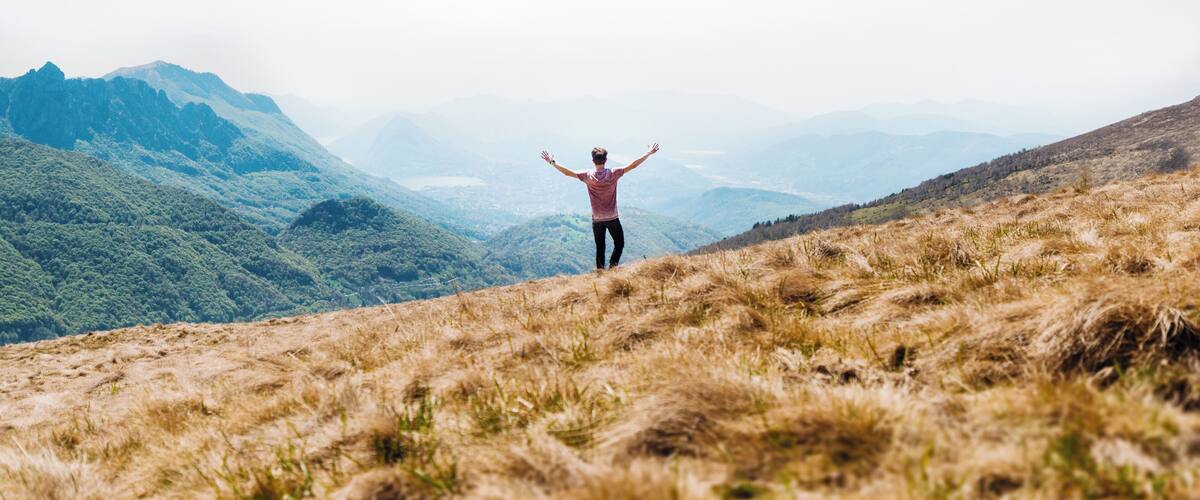 A person standing on a grassy hill with arms raised, overlooking a mountainous landscape.