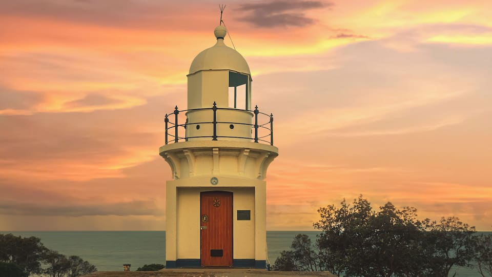 Un phare avec une porte en bois, entouré d’arbres, se dressant sur fond de ciel au coucher du soleil.