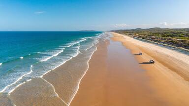 A wide sandy beach with gentle waves, a clear blue sky, and distant hills.