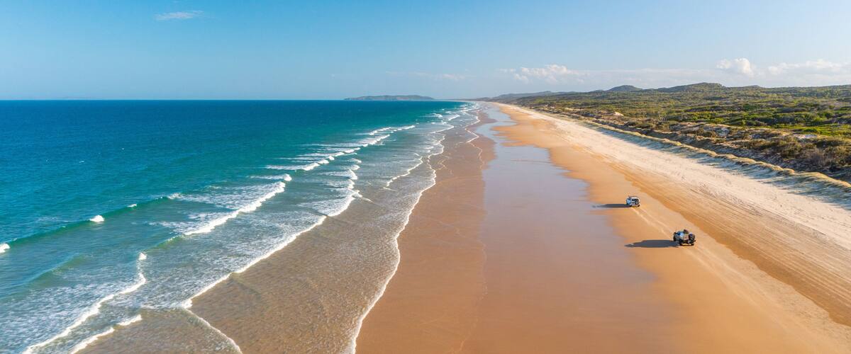 A wide sandy beach with gentle waves, a clear blue sky, and distant hills.