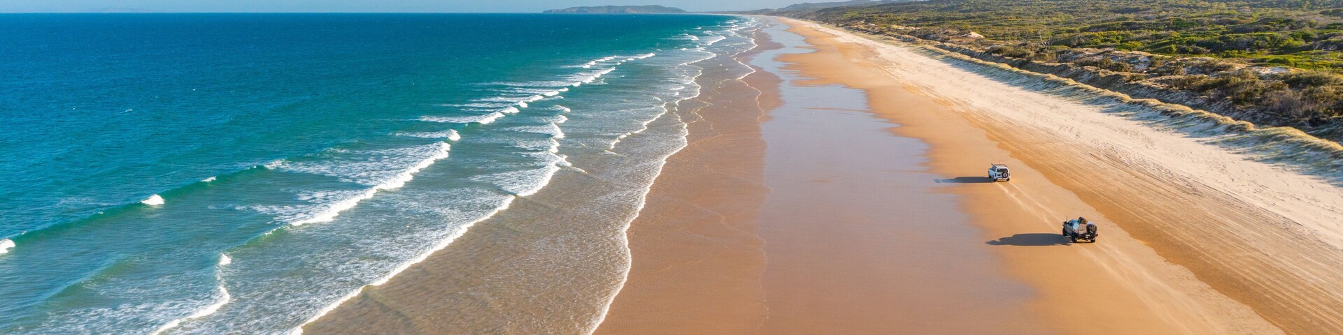 A wide sandy beach with gentle waves, a clear blue sky, and distant hills.