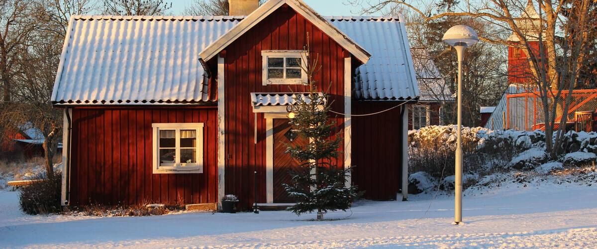 A red wooden house with a snow-covered roof and a small tree in front.