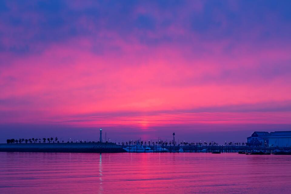Ein Sonnenuntergang über einem Wasserufer mit Booten, einem Leuchtturm und Gebäuden am Ufer.