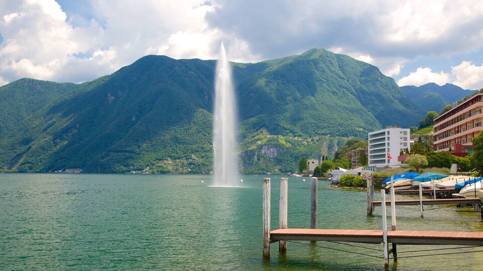 A lake with a fountain, mountains, and buildings.