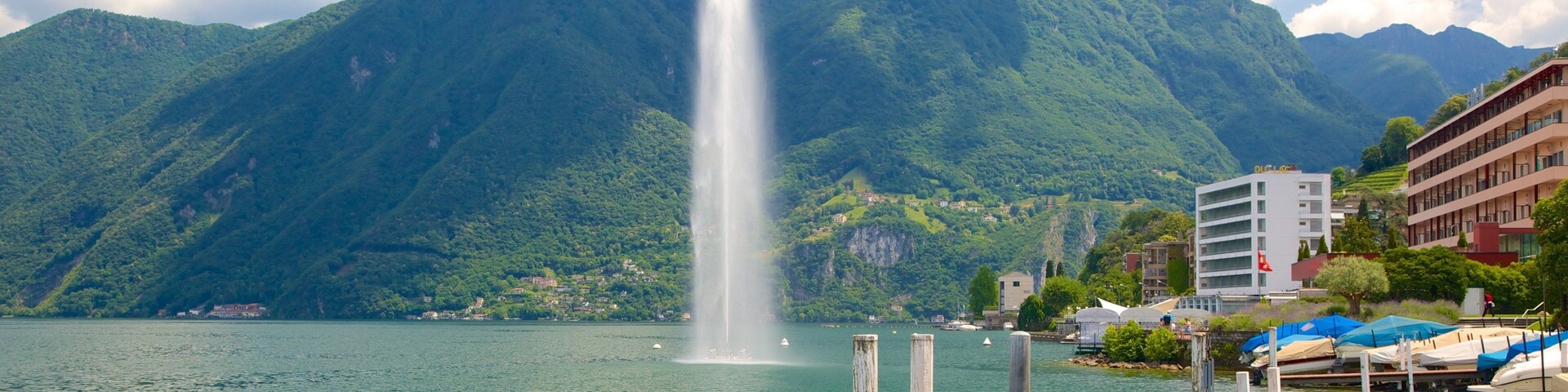 A lake with a fountain, mountains, and buildings.