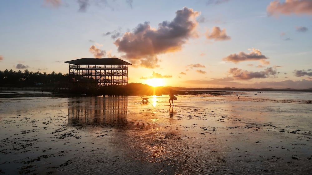 A beach at sunset with a gazebo structure and a person with a surfboard.