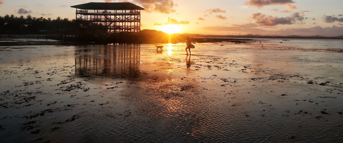 A beach at sunset with a gazebo structure and a person with a surfboard.