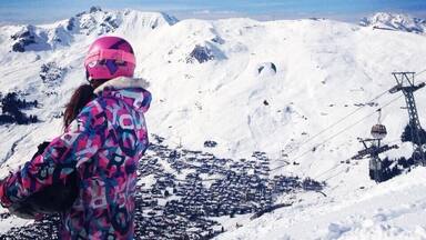 A person in a colorful jacket and pink helmet overlooking a snow-covered mountain range with a ski lift in the background.