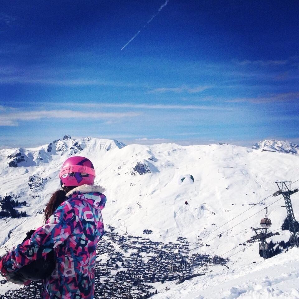 A person in a colorful jacket and pink helmet overlooking a snow-covered mountain range with a ski lift in the background.