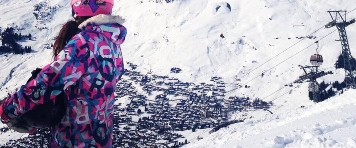 A person in a colorful jacket and pink helmet overlooking a snow-covered mountain range with a ski lift in the background.