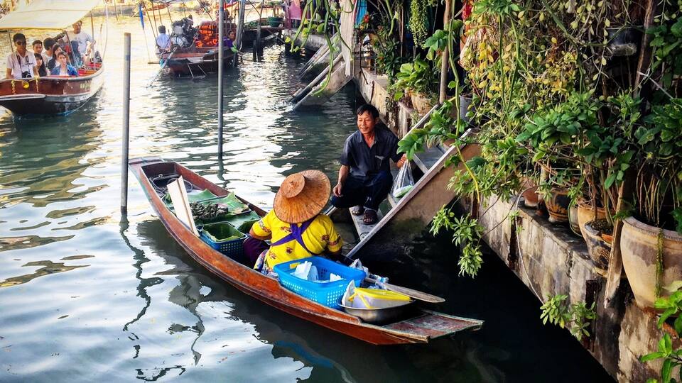 A narrow waterway with boats, a person in a boat wearing a straw hat, and a building with colorful flags in the background.