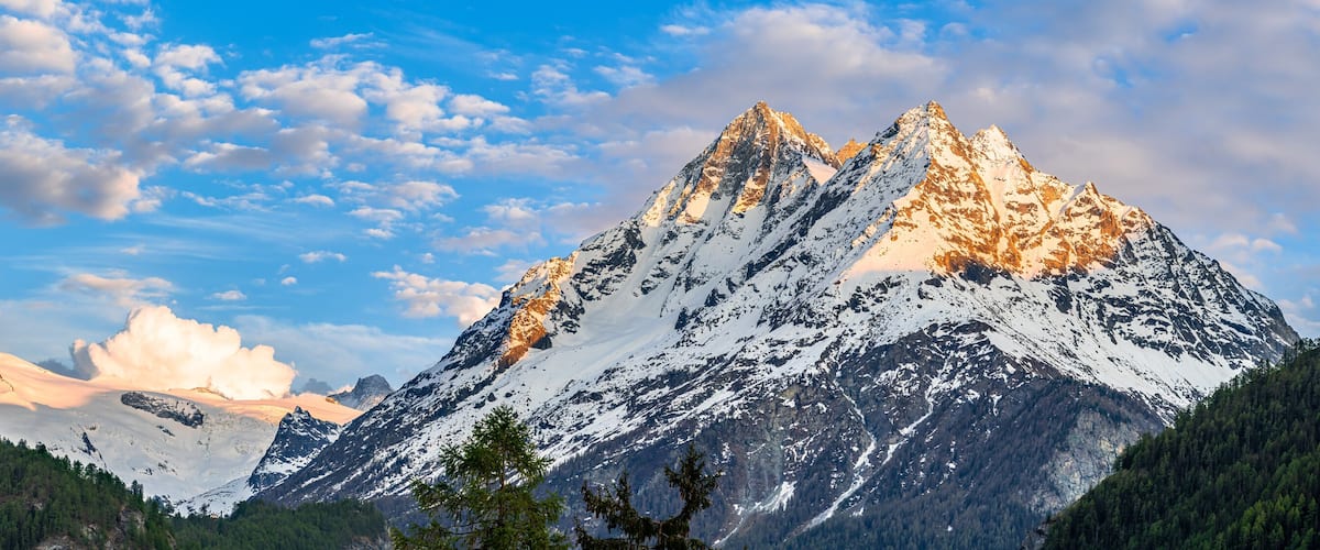 A mountain range with snow-capped peaks, dense forests, and a clear blue sky.