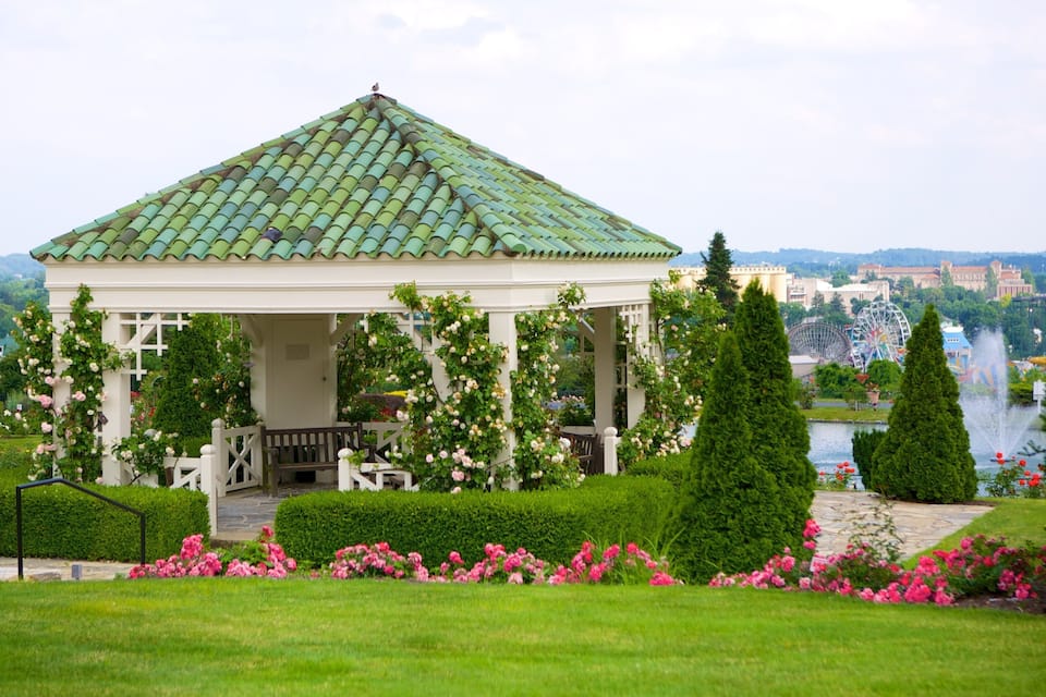 A gazebo with a green roof surrounded by lush gardens and a lake.