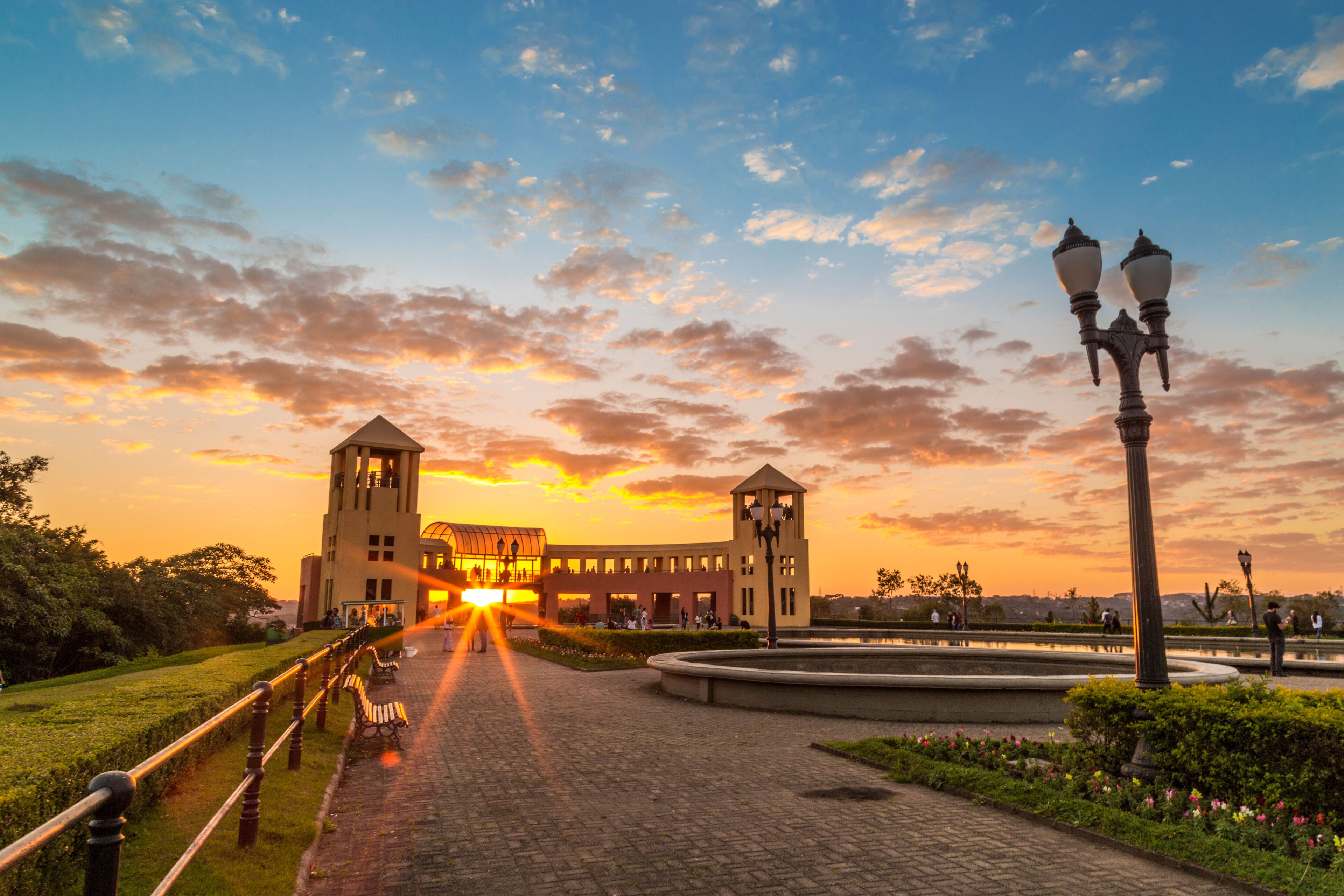 Um pôr do sol sobre um parque com um edifício, uma fonte e um caminho.