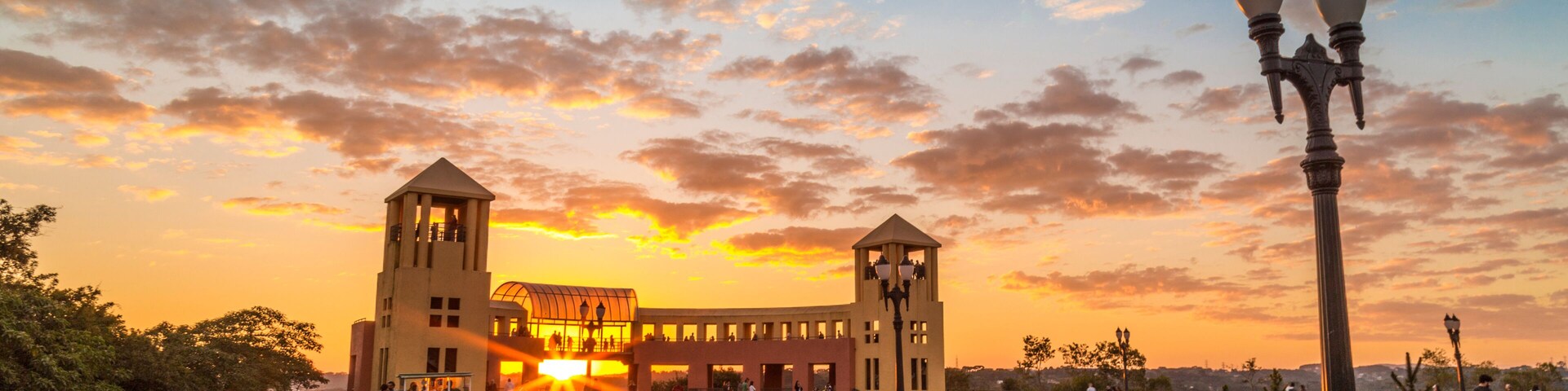 Um pôr do sol sobre um parque com um edifício, uma fonte e um caminho.