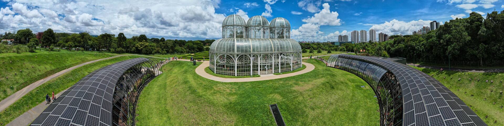 A large greenhouse with multiple domes, surrounded by green fields and solar panels.