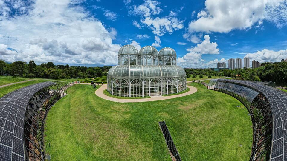 A large greenhouse with multiple domes, surrounded by green fields and solar panels.