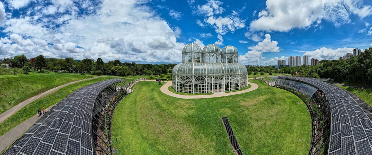 A large greenhouse with multiple domes, surrounded by green fields and solar panels.