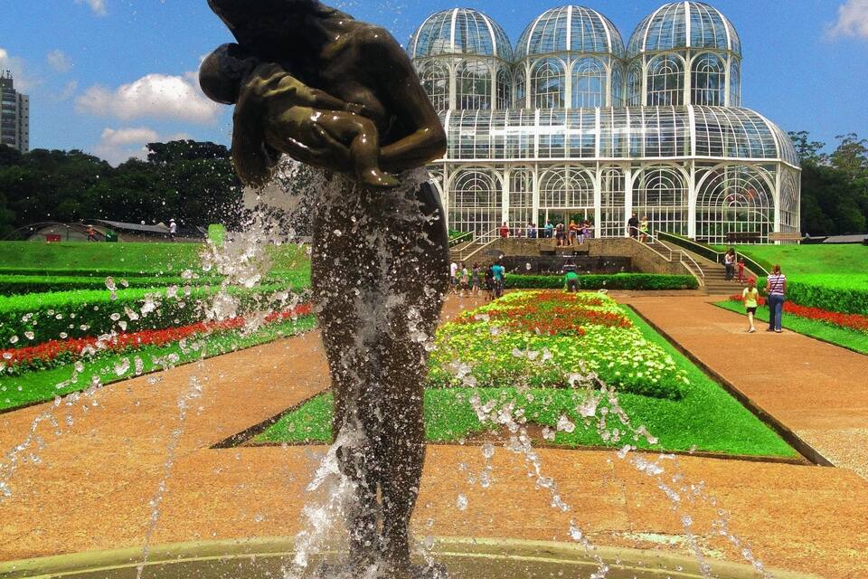 A fountain sculpture in a park with a greenhouse in the background.