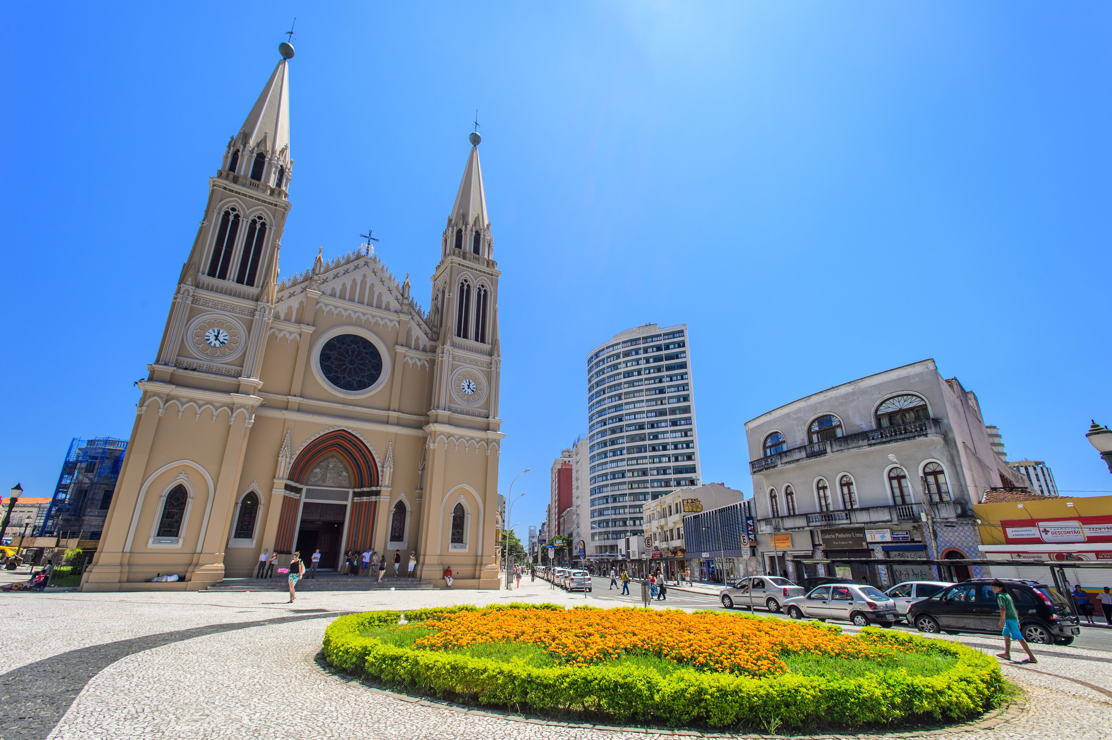 A historic church with twin spires, a modern high-rise building, and a flower garden in a city square.