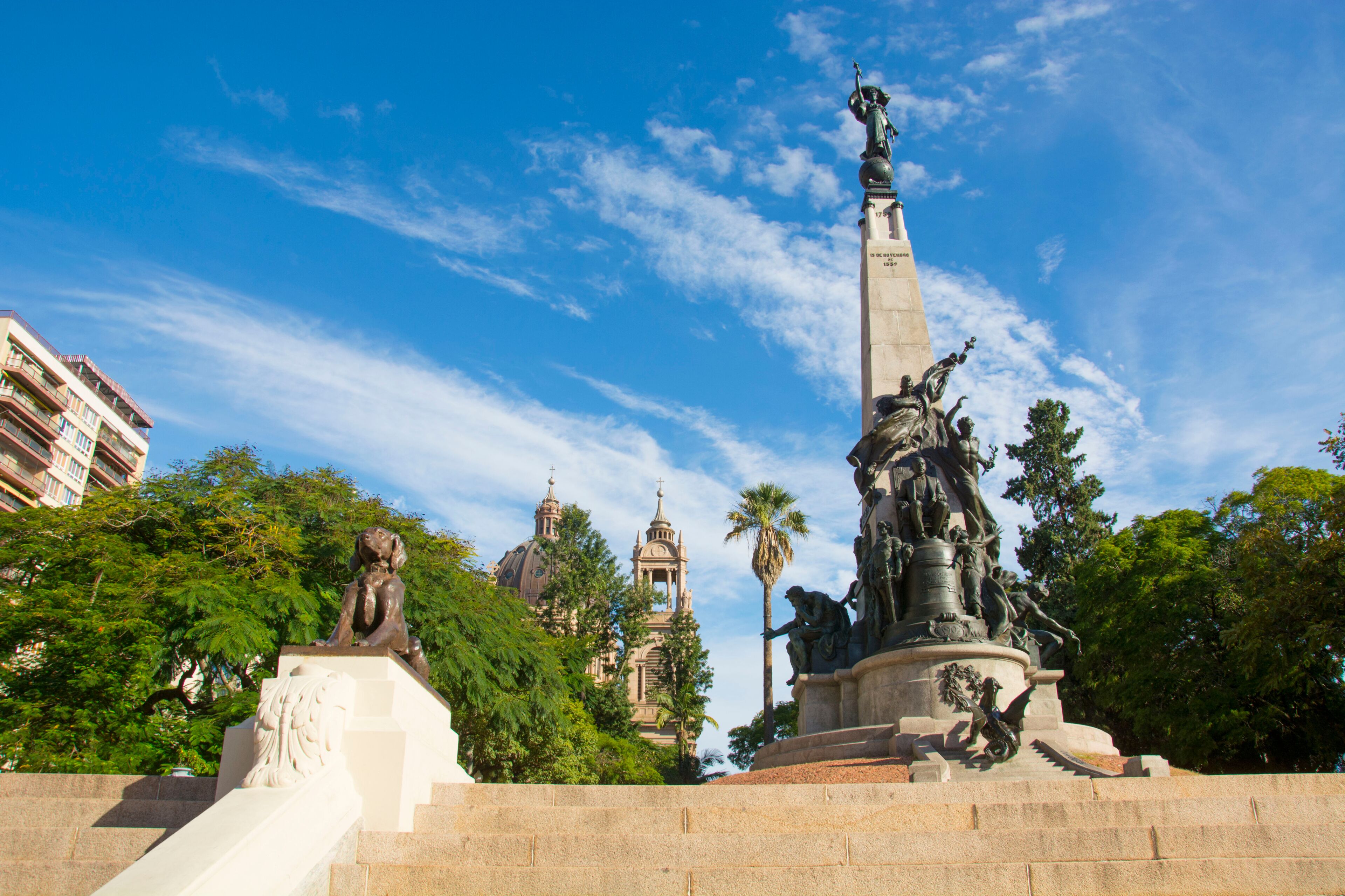 A monument with a statue, surrounded by trees and a historic building with domes in the background.