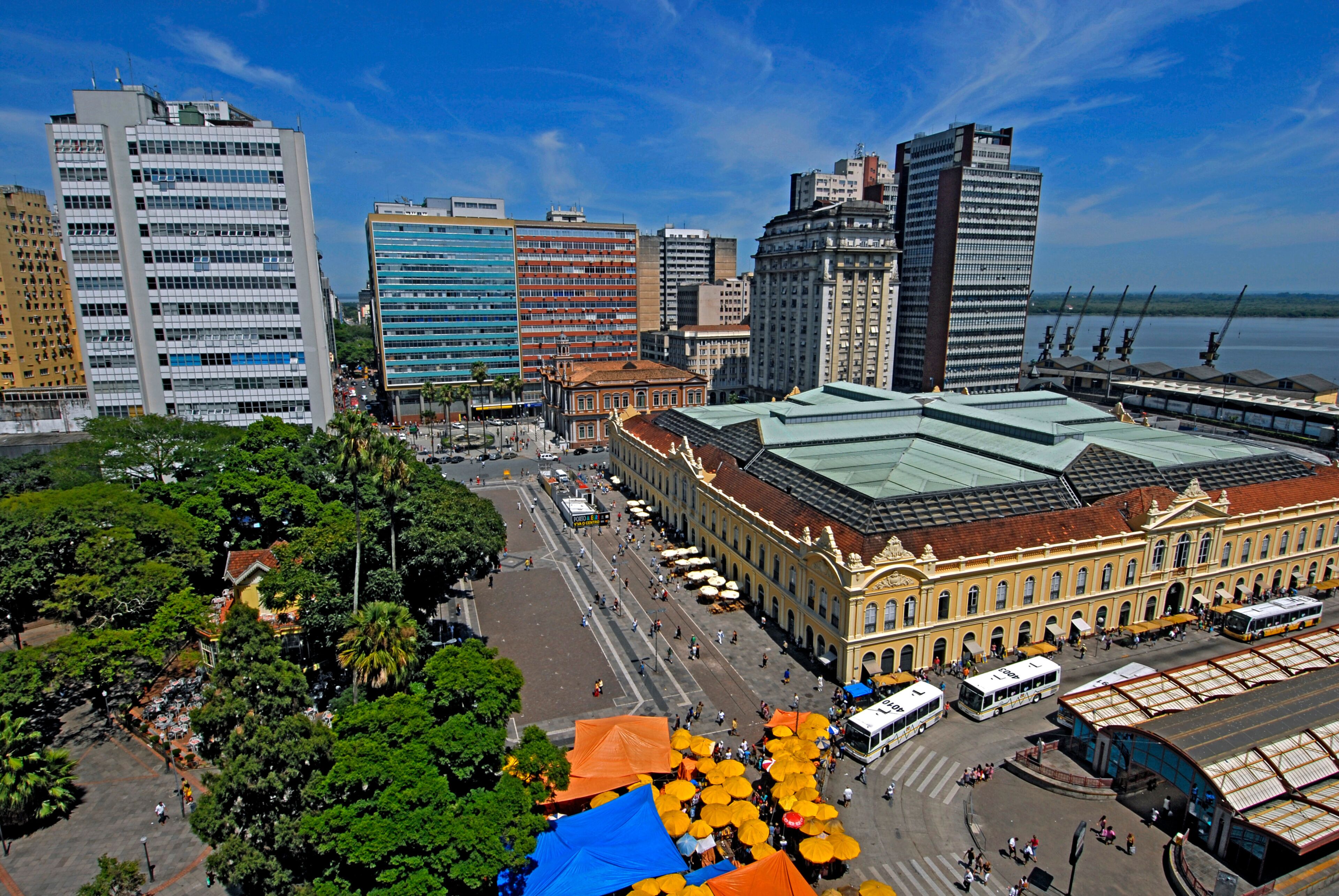A cityscape with a mix of modern and historic buildings, a central plaza with a large covered structure, and a waterfront in the background.