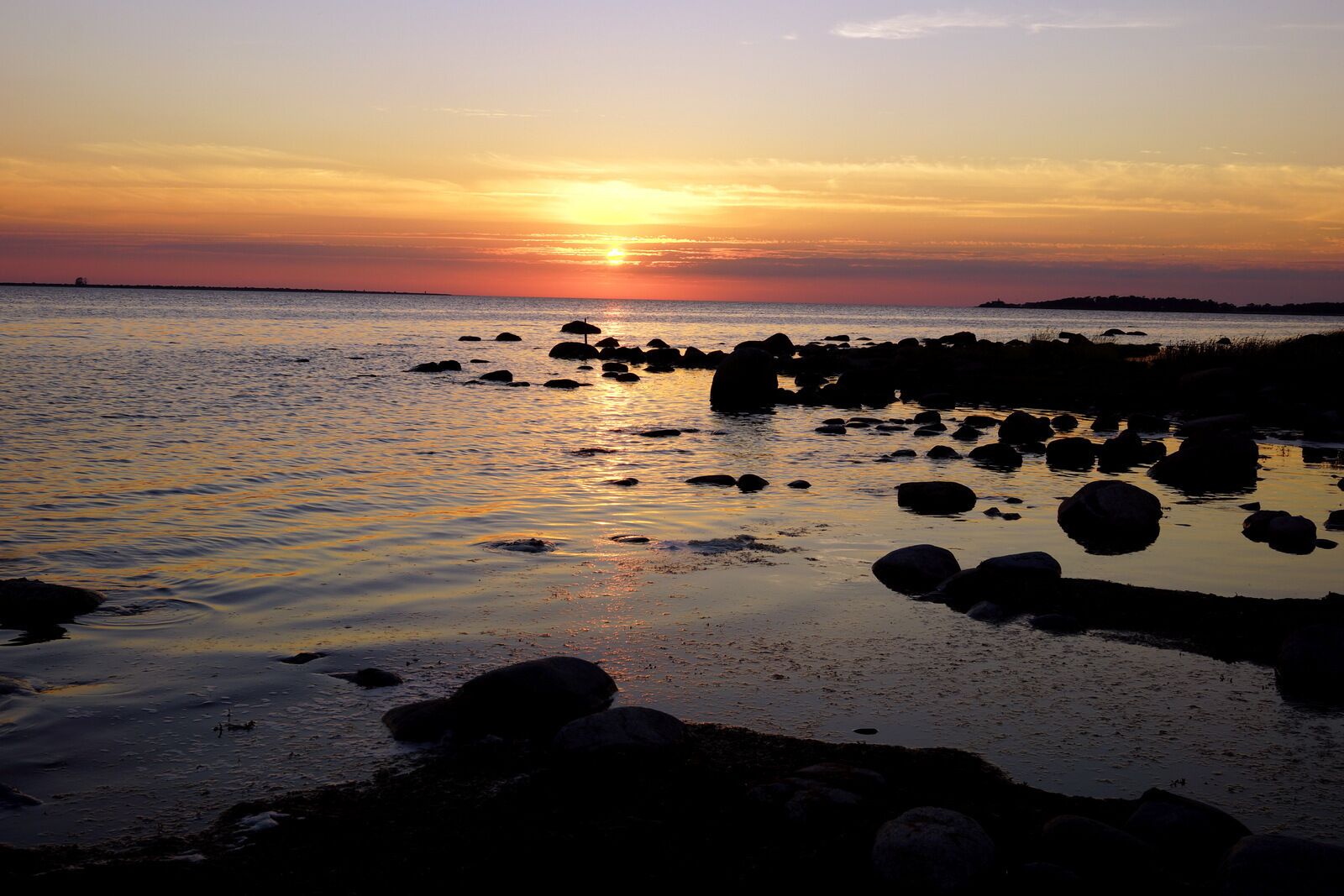 A sunset over a rocky shoreline with calm water reflecting the sky.