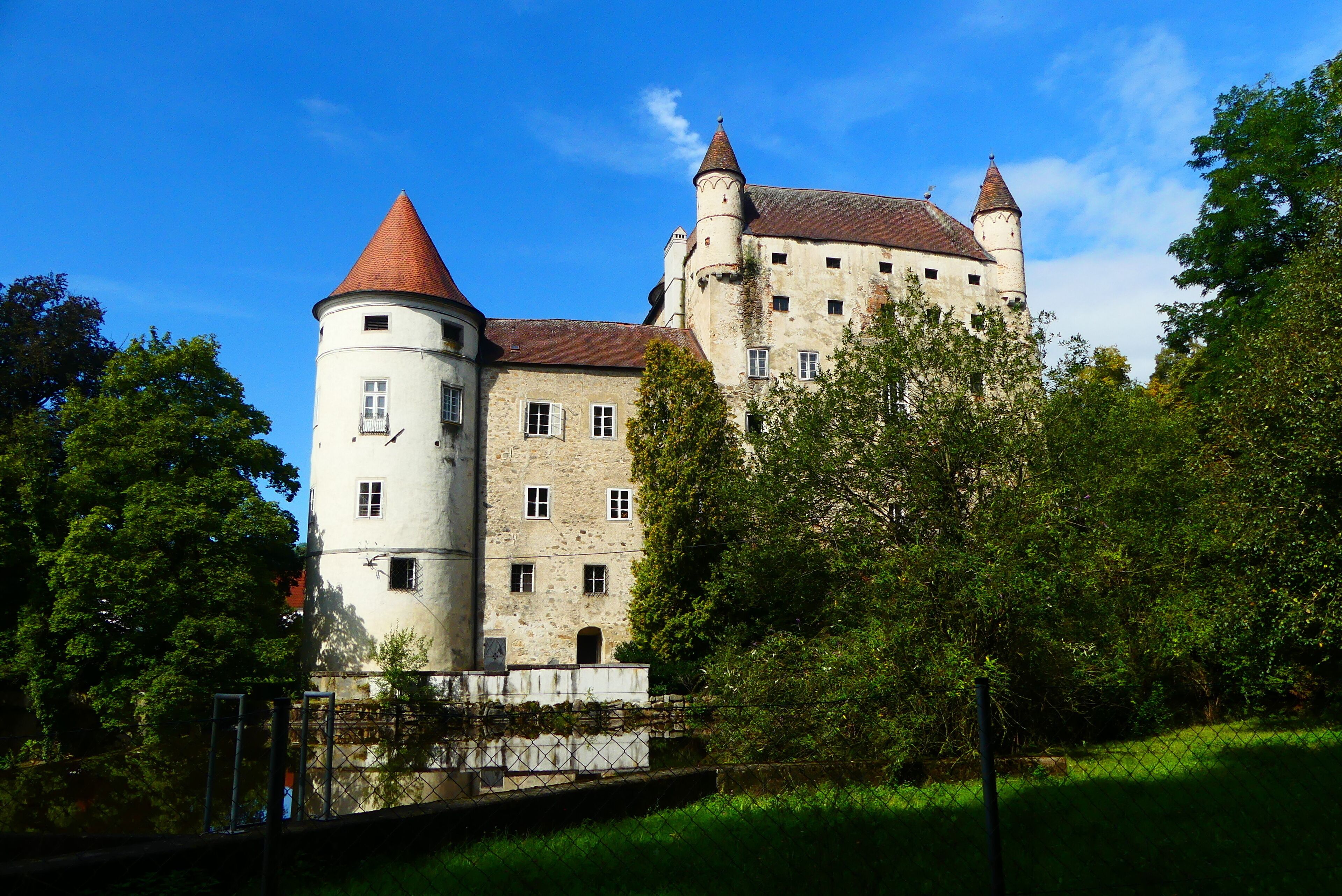 A historic castle with a round tower and a conical roof, surrounded by lush greenery.