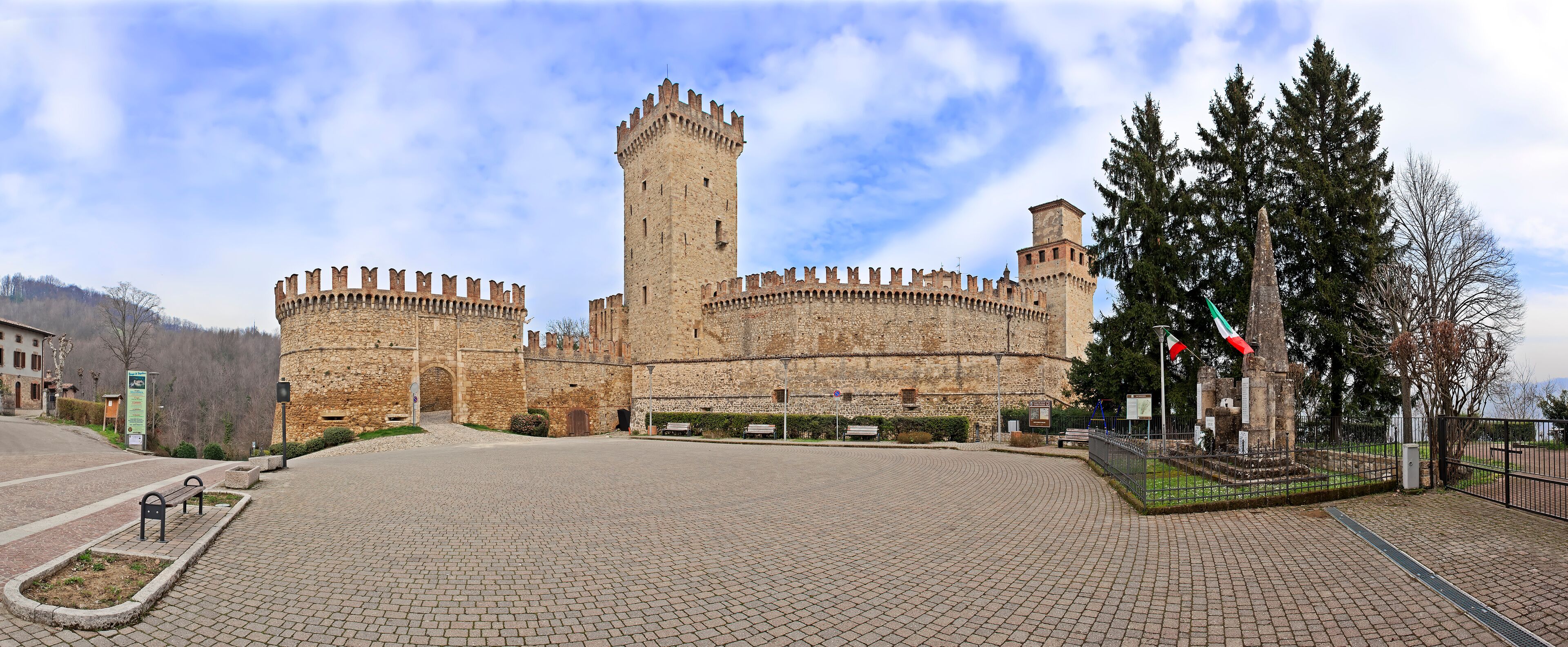 A historic castle with a tall central tower, surrounded by a paved courtyard and a flagpole with flags.