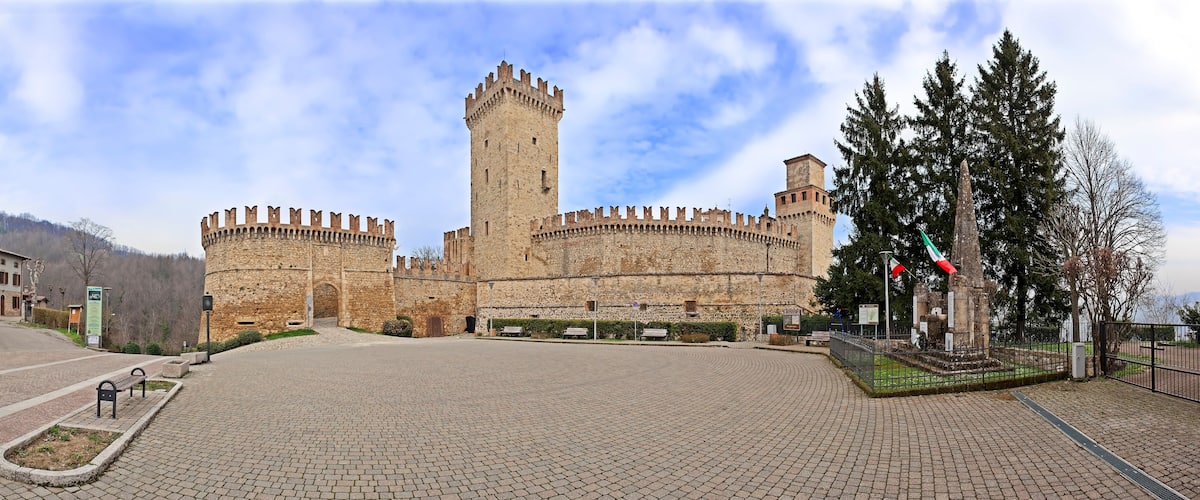 A historic castle with a tall central tower, surrounded by a paved courtyard and a flagpole with flags.