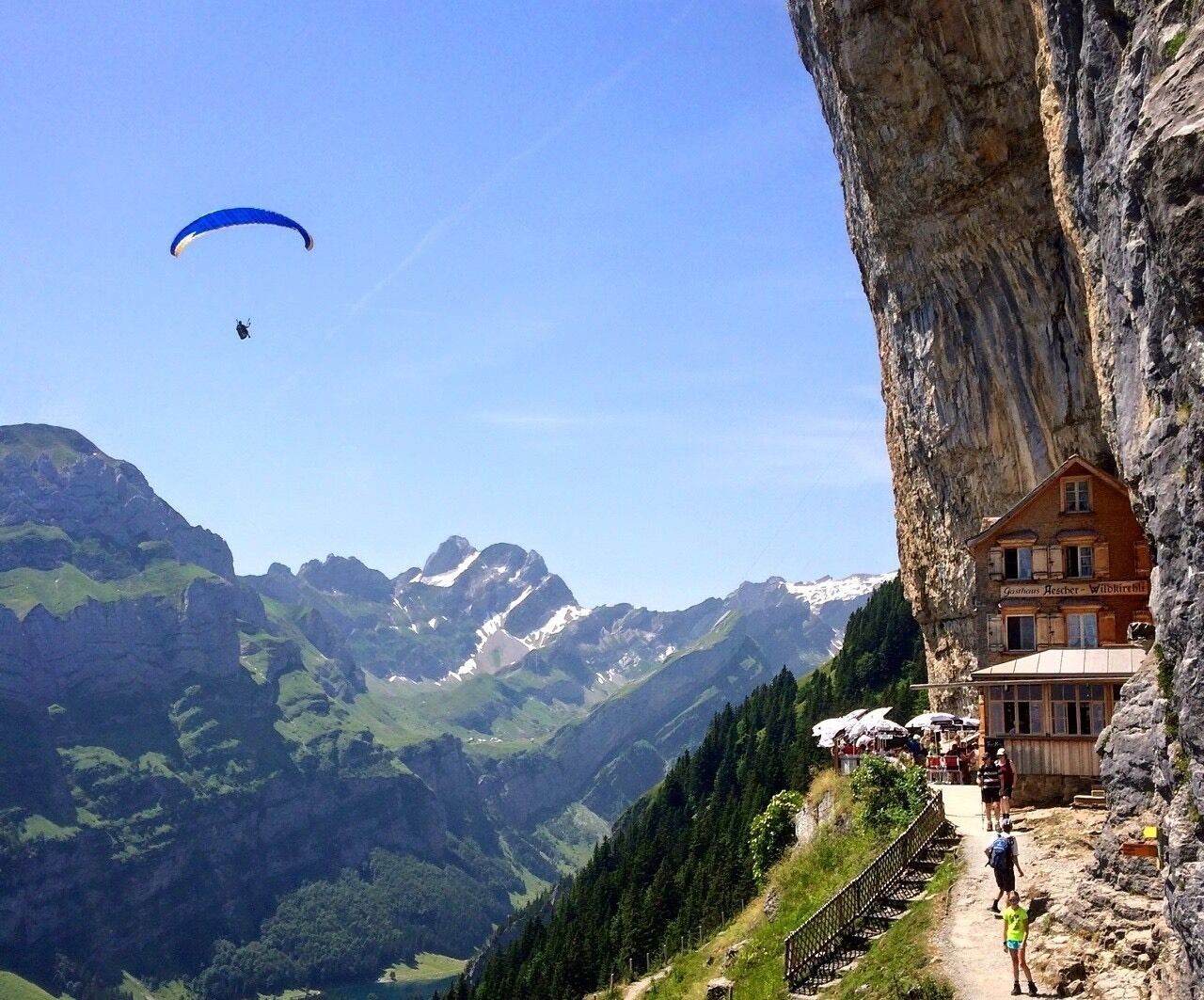 Una persona practicando parapente sobre un paisaje montañoso con una cabaña en el borde de un acantilado.