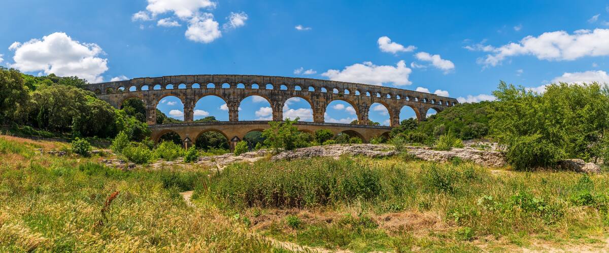 A large stone aqueduct with multiple arches, surrounded by lush greenery and a clear blue sky.