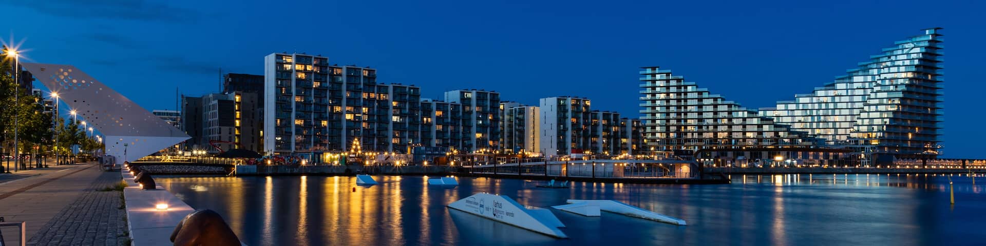 A waterfront cityscape at dusk with illuminated buildings and a calm river.