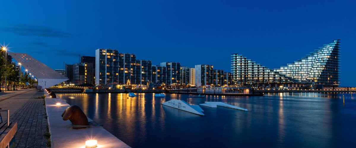 A waterfront cityscape at dusk with illuminated buildings and a calm river.