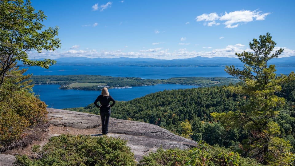 A person standing on a rocky outcrop overlooking a vast lake surrounded by dense forest and distant mountains.