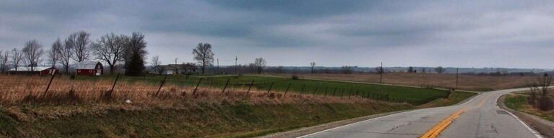 A rural road with yellow centerline markings, surrounded by open fields and a cloudy sky.