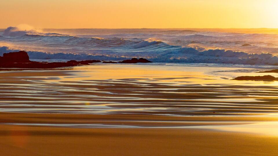 A beach with waves crashing onto the shore and a sunset in the background.