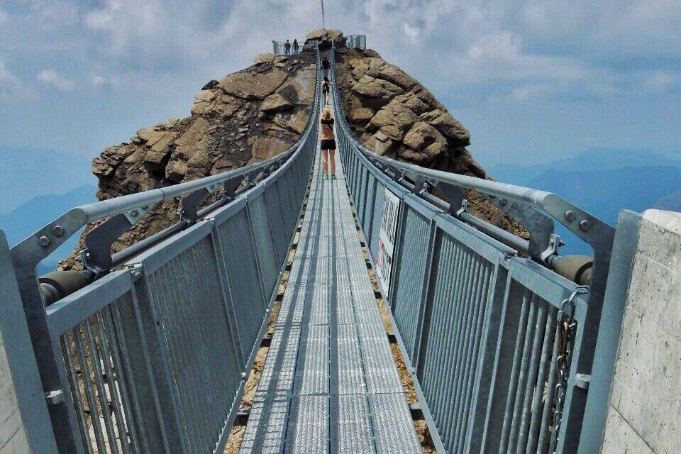 A metal bridge with a metal railing, leading to a rocky peak with a red flag.